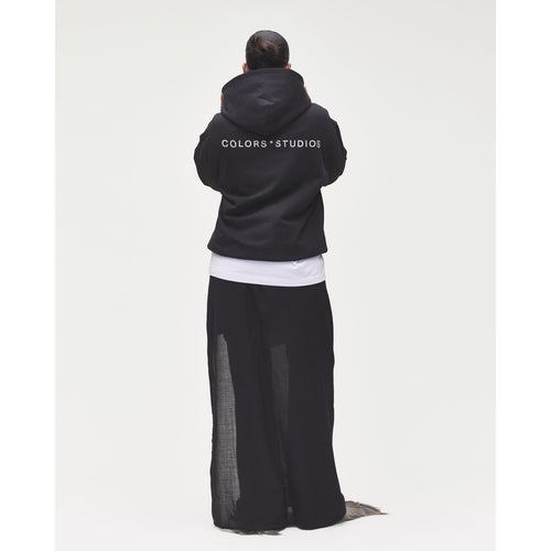 Rückansicht einer Person mit schwarzem COLORS*STUDIOS Hoodie und großem weißen Logo-Print auf dem Rücken, kombiniert mit einer schwarzen Wide-Leg-Hose vor hellem Studiohintergrund.