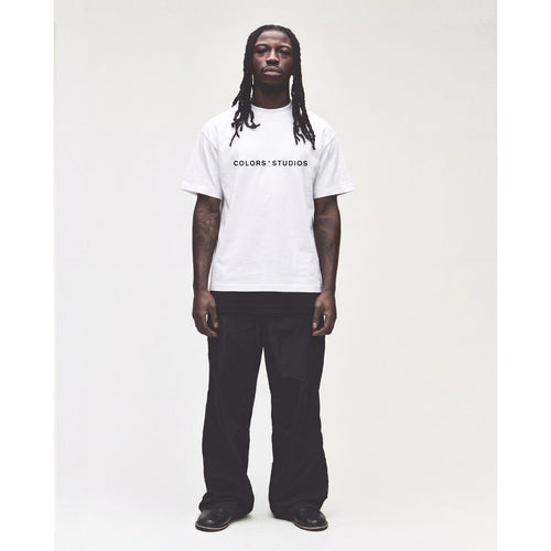 Mann trägt ein weißes COLORS*STUDIOS T-Shirt mit schwarzem Logo-Print auf der Brust, kombiniert mit einer schwarzen Wide-Leg-Hose. Studioaufnahme vor hellem Hintergrund.