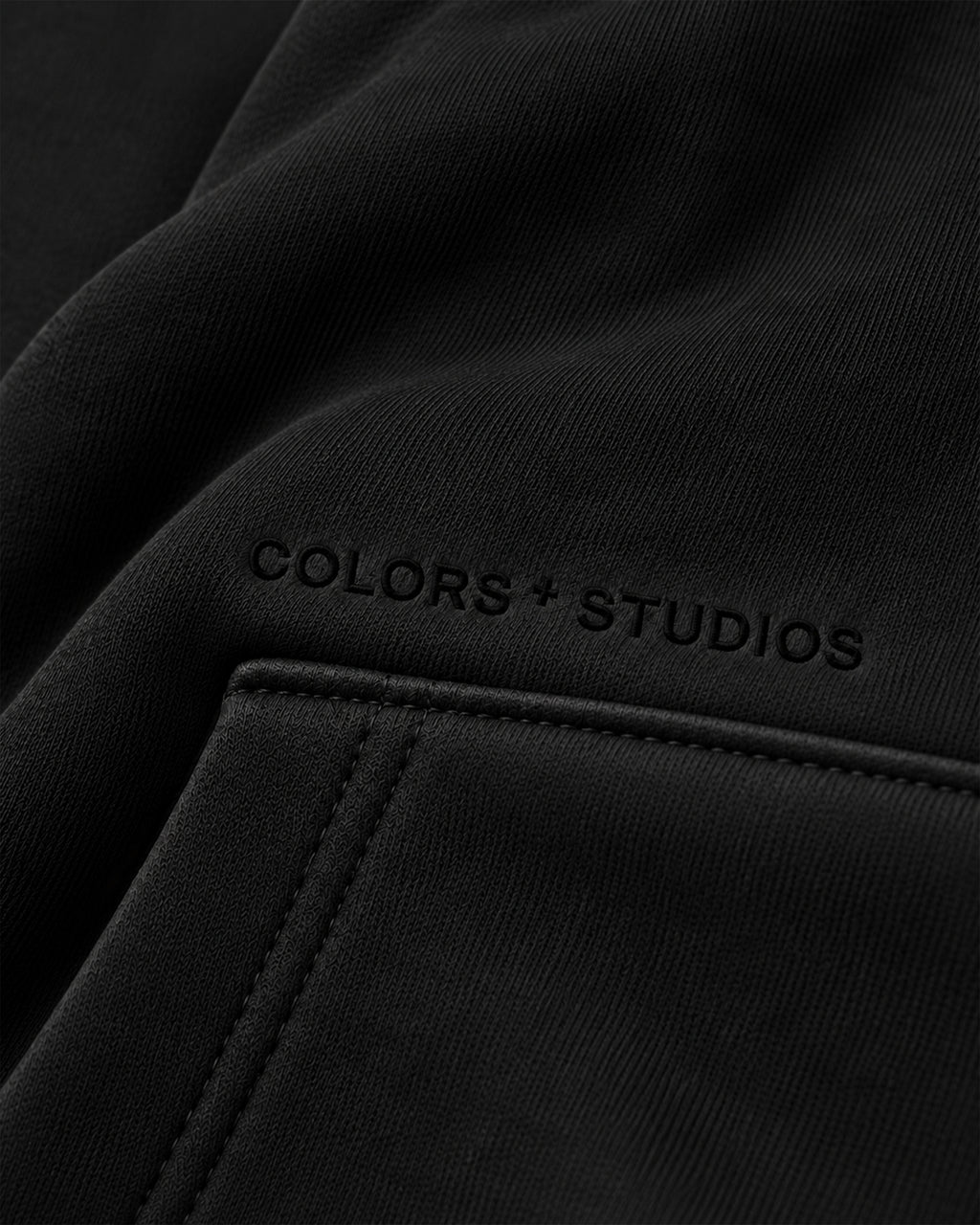 Close-up of black hoodie fabric and kangaroo pocket with subtle tonal COLORS+STUDIOS logo detail.