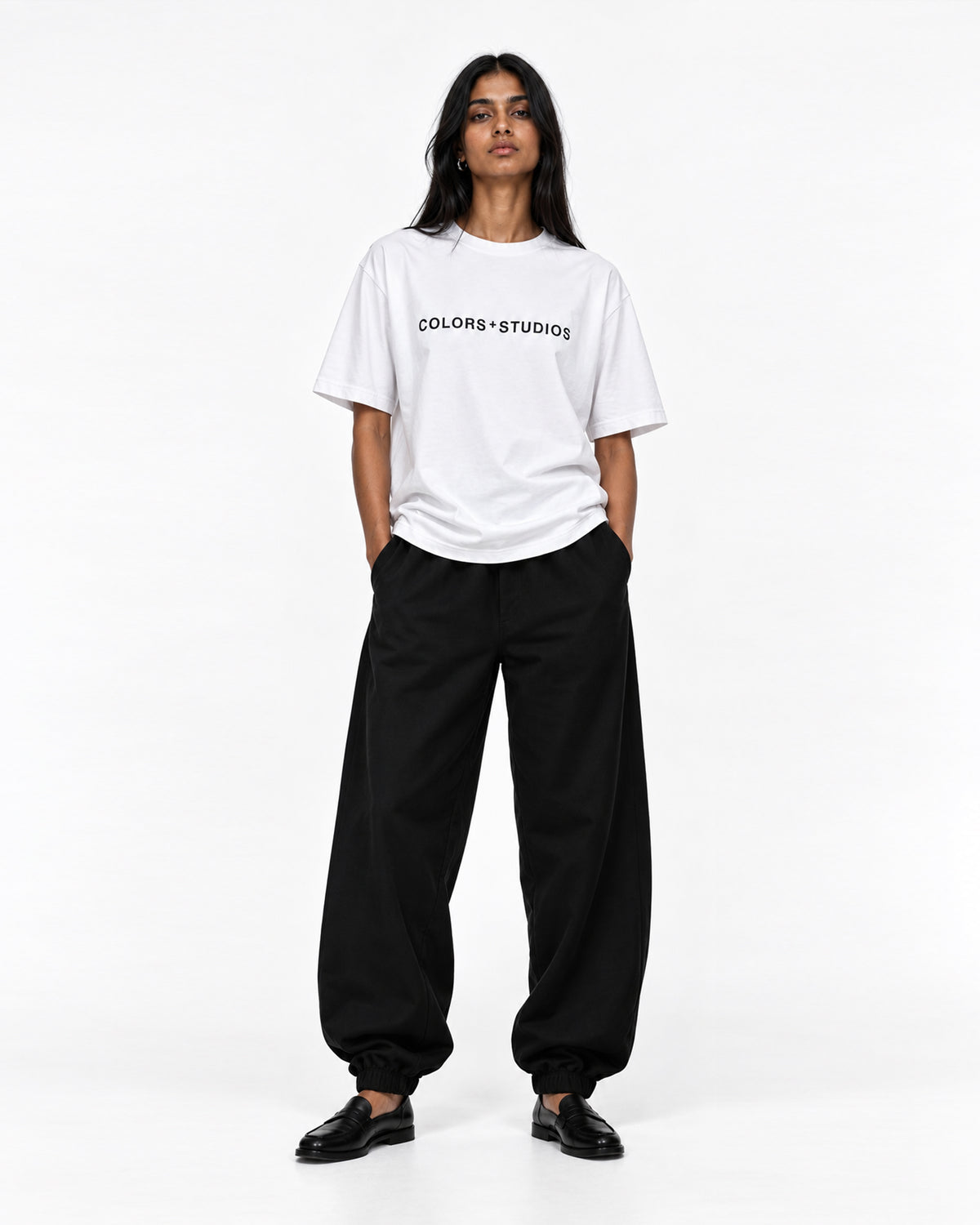 Front view of model wearing a white T-shirt with black COLORS+STUDIOS chest logo, styled with loose black trousers.