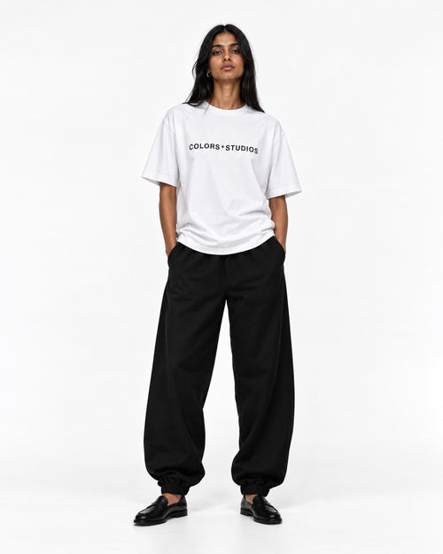 Front view of model wearing a white T-shirt with black COLORS+STUDIOS chest logo, styled with loose black trousers.