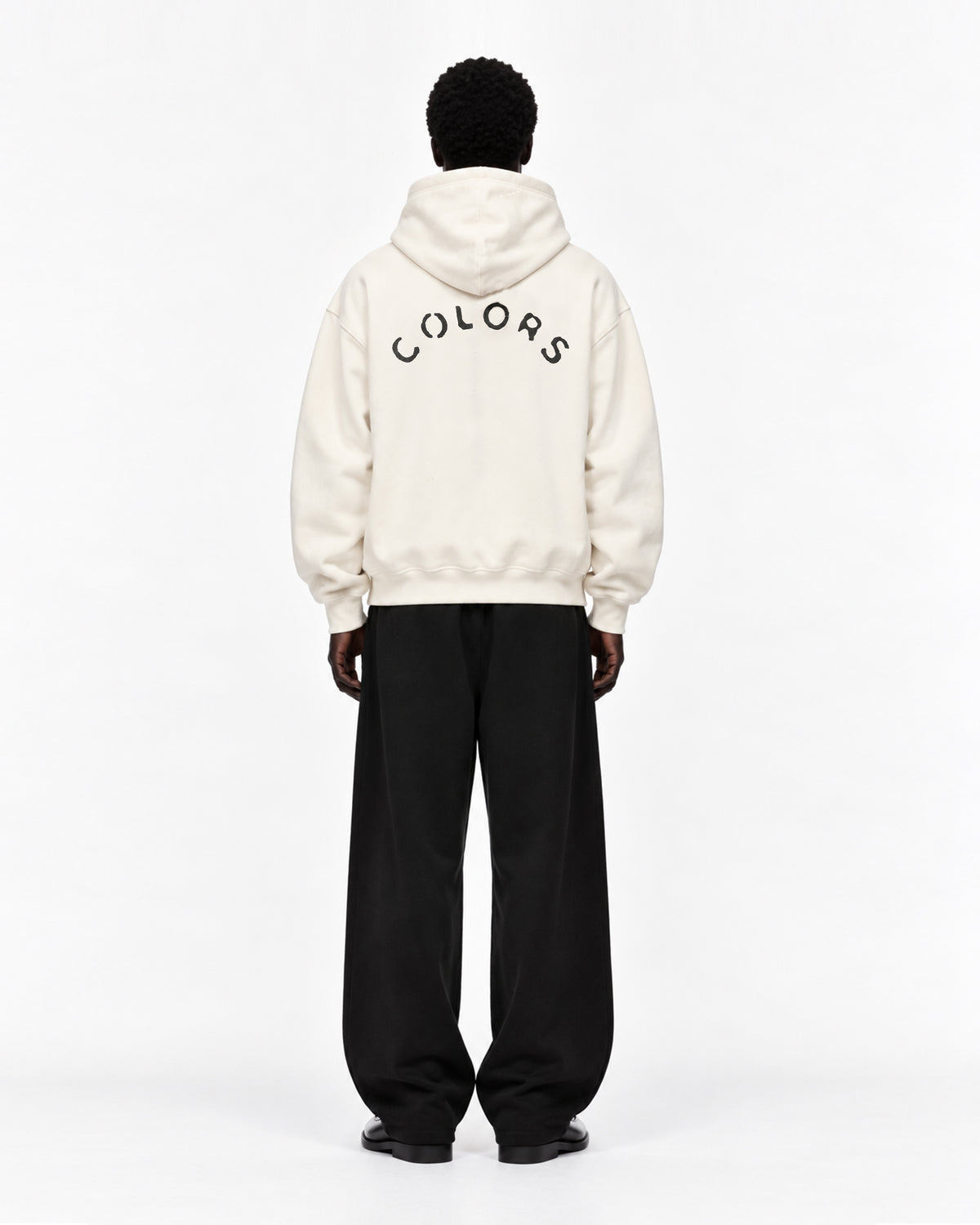 Rückansicht eines Models in cremeweißem Oversized-Hoodie mit schwarzem, bogenförmigem „COLORS“-Schriftzug auf dem Rücken, dazu weite schwarze Hose und schwarze Schuhe