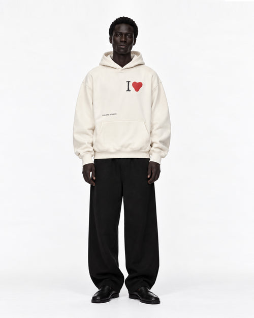 Model in cremeweißem Oversized-Hoodie mit „I ♥“-Print auf der Brust und kleinem Schriftzug, dazu weite schwarze Hose und schwarze Loafer, Vorderansicht