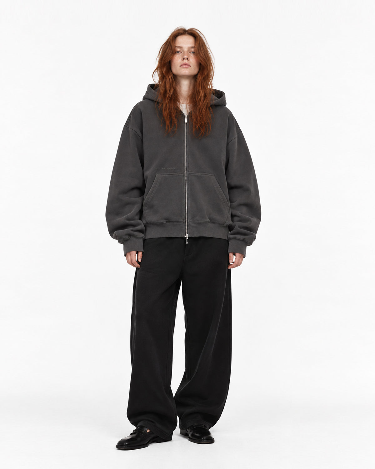 Model mit roten Haaren in anthrazitfarbener Oversized-Zip-Hoodie mit silbernem Reißverschluss, dazu weite schwarze Hose und schwarze Loafer, Vorderansicht