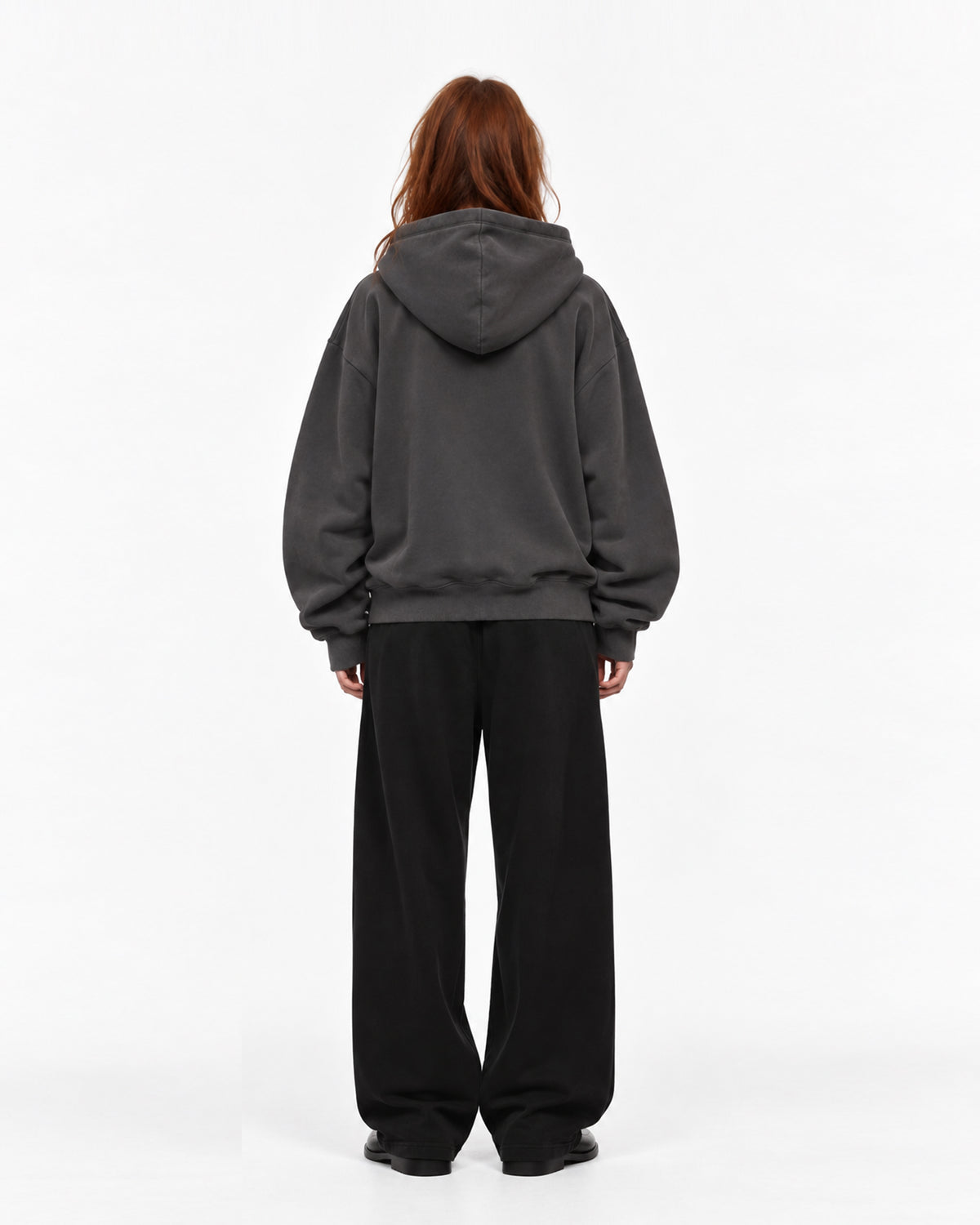 Rückansicht eines Models mit roten Haaren in anthrazitfarbenem Oversized-Hoodie mit Kapuze, dazu weite schwarze Hose