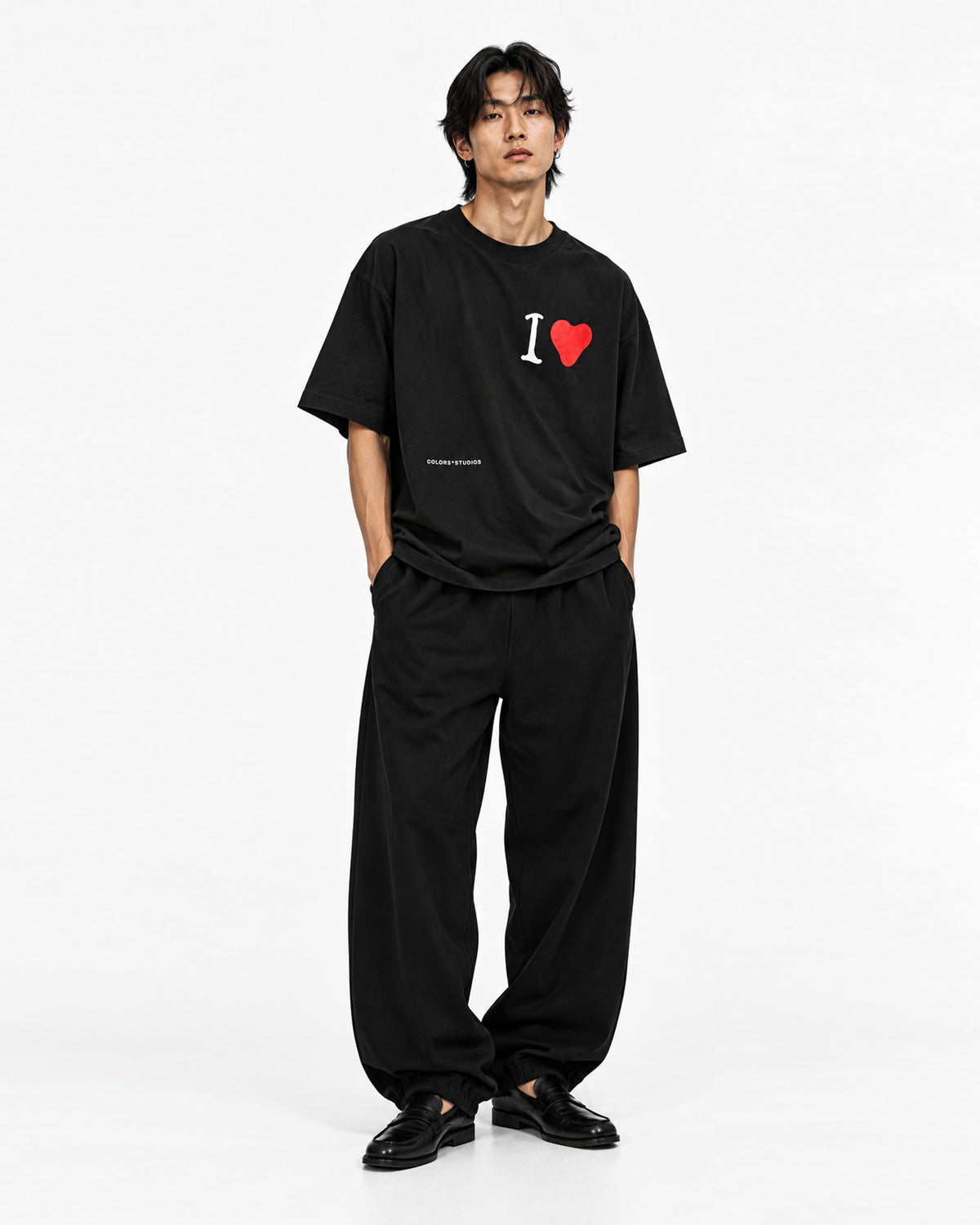 Model wearing an oversized black T-shirt with white “I” and red heart chest graphic, styled with loose black trousers.